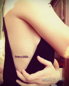 Фото тату: надпись "Live a little" на плече девушки. Небольшая, но вдохновляющая татуировка, выполненная черными чернилами.