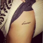 Фото тату: небольшая надпись "dreamer" (мечтатель) на английском языке, выполненная тонким шрифтом, расположена на боку. Минималистичный и вдохновляющий рисунок.