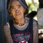 Фотография пожилой женщины с традиционными татуировками на лице и руках, демонстрирующими культурное наследие.