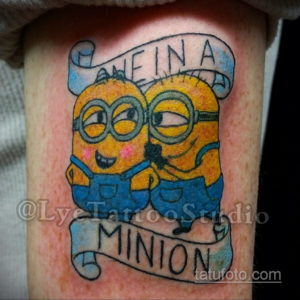Фото тату на руке: Миньон в рамке с надписью "MINION", яркие цвета, мультяшный стиль, синий фон.