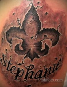 Черно-серая татуировка флёр-де-лис с именем "Stephanie" внизу. Эффект потертости и трещин придает рисунку винтажный вид.