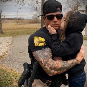 Фотография шерифа с татуировками на руках, обнимающего девушку на фоне сельской местности.
