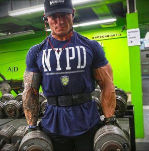 Фотография полицейского в спортивной форме с татуировками на руках, занимающегося в спортзале с гантелями.
