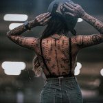 Фото девушки с множеством татуировок на спине, стоящей спиной к камере. Татуировки включают в себя геометрические узоры, иероглифы и изображения животных, выполненные в черном цвете.