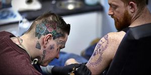 Мастер тату делает татуировку клиенту на плече. Видны татуировки на лице и шее мастера. Яркие цвета и традиционный стиль.
