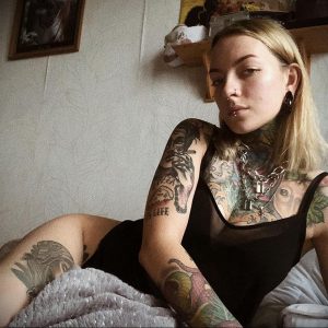 Фото девушки с множеством татуировок на руках, шее и плечах, а также с пирсингом на губе и ушах. Татуировки выполнены в различных стилях и цветах.