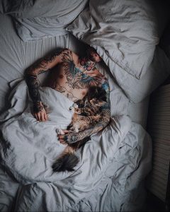 Фото татуированного тела девушки, лежащей в постели с кошкой. Татуировки в стиле олдскул и акварель, создающие атмосферу свободы и любви.