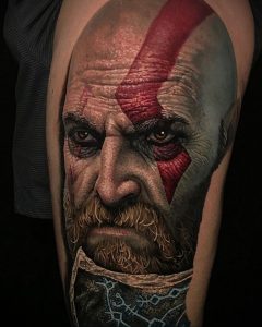Фото тату на руке: реалистичный портрет Кратоса из игры God of War. Детализированная татуировка с красными татуировками на лице и бородой.