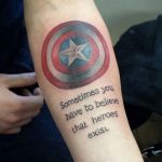 Фото тату щита Капитана Америки с надписью "Sometimes you have to believe that heroes exist" на руке. Классический стиль, четкие линии.