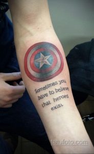 Фото тату щита Капитана Америки с надписью "Sometimes you have to believe that heroes exist" на руке. Классический стиль, четкие линии.