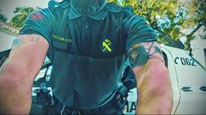 Фотография сотрудника испанской Guardia Civil с татуировками на руках, сидящего за рулем автомобиля. Он одет в форму и выглядит сосредоточенно.