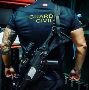 Фотография сотрудника испанской Guardia Civil с татуировками на руках, держащего оружие. Он одет в форму и выглядит серьезно.