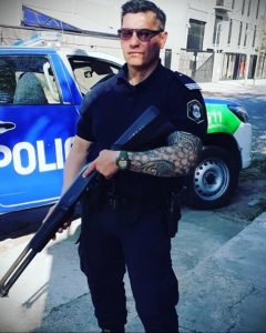 Фотография полицейского в форме с татуировкой на предплечье, держащего оружие, демонстрирующая силу и готовность к действию.