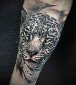 Фото тату: реалистичный рисунок головы леопарда с желтыми глазами в черно-серых тонах на предплечье, детализированная проработка шерсти.