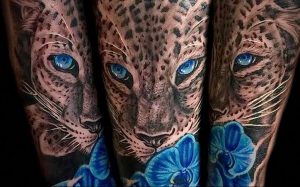 Фото реалистичной татуировки гепарда на руке. Татуировка выполнена в черно-серых тонах с яркими голубыми глазами и голубыми цветами.