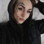 Фото тату: Черная татуировка надписи 'Delirious' на лбу девушки. Дополняется небольшими татуировками на лице.