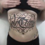 Фото тату: стильная надпись на животе, выполненная черными чернилами в каллиграфическом стиле. Изящные завитки обрамляют текст, создавая эффектный рисунок.