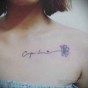 Фото тату надписи 'Carpe Diem' с маленьким синим цветком на ключице. Изящный шрифт и нежные цвета.