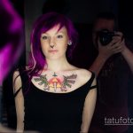 Фотография девушки с яркой татуировкой на груди в виде крыльев, окружающих сердце. Девушка с фиолетовыми волосами позирует на фоне размытого фона.