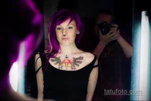 Фотография девушки с яркой татуировкой на груди в виде крыльев, окружающих сердце. Девушка с фиолетовыми волосами позирует на фоне размытого фона.