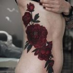 Фото тату на боку с изображением красных пионов. Реалистичный рисунок с детальной проработкой лепестков и листьев. Идеальный эскиз тату для женщин.