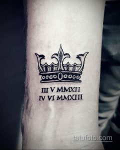 Фото тату на предплечье: черная корона с римскими цифрами под ней. Стильный и значимый рисунок, возможно, символизирующий важные даты.