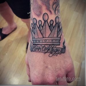 Фото тату: черно-серая татуировка короны с лентой и надписью 'Born to Reign', расположенная на руке.