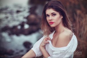 Фото красивой девушки с татуировкой на плече, смотрящей в камеру. Девушка одета в белую рубашку.