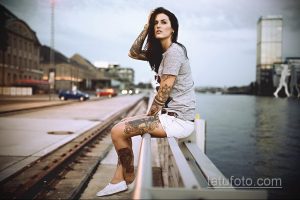 Фото стильной девушки, сидящей на парапете с татуировками на руках и ногах. На заднем плане виднеется городская архитектура.