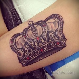 Фото тату большой короны с красными камнями на руке. Детализированный рисунок татуировки, выполненный в реалистичном стиле, выглядит эффектно и богато.