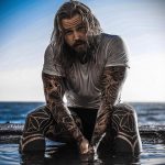Фотография мужчины с татуировками на теле, сидящего на камнях у моря. Идеально для поиска идей для тату в стиле геометрия и трайбл.