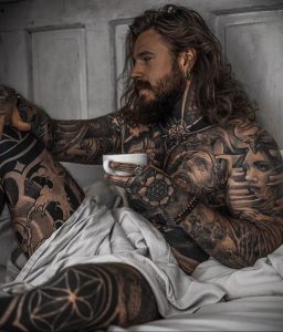 Фотография мужчины с татуировками на теле, сидящего в постели с чашкой кофе. Идеально для поиска идей для тату в стиле реализм и флористика.