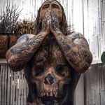 Фотография мужчины с огромной татуировкой черепа на груди и животе, стоящего под струями воды. Идеально для поиска идей для тату в стиле реализм.