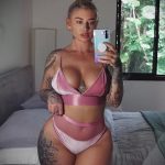 Фотография девушки в розовом белье, демонстрирующей татуировки на руках и бедрах. Девушка делает селфи в спальне.