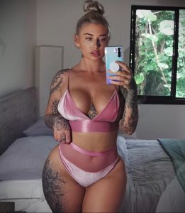 Фотография девушки в розовом белье, демонстрирующей татуировки на руках и бедрах. Девушка делает селфи в спальне.