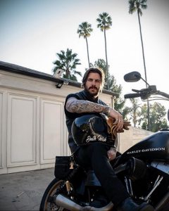 Фотография мужчины с бородой, сидящего на черном мотоцикле Harley-Davidson, демонстрирующего татуировки на руке.