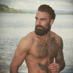 Фотография мужчины с татуировками на груди и руках, стоящего у воды с задумчивым видом.
