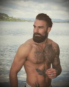 Фотография мужчины с татуировками на груди и руках, стоящего у воды с задумчивым видом.