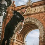 Фотография мужчины с татуировкой на предплечье, стоящего на фоне красивой архитектуры города.