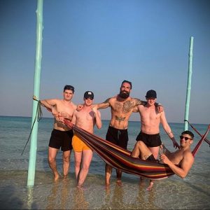 Фотография группы мужчин на пляже, один из которых демонстрирует татуировки на груди и руках.