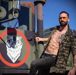 Мужчина в военной куртке с татуировкой волка на груди позирует на фоне бронетранспортера под ярким солнцем. Фотография подчеркивает силу и мужественность.