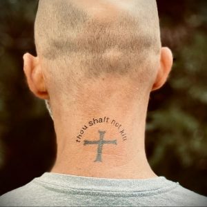 Фото тату креста с надписью 'thou shalt not kill' на затылке мужчины. Минималистичная татуировка в серо-черных тонах, выполненная аккуратным шрифтом.