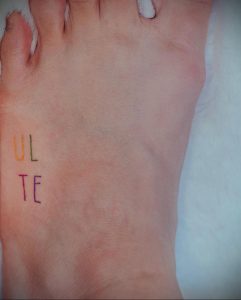 Фото тату: надпись 'ULTE' выгравирована на верхней части стопы, используя разные цвета чернил (оранжевый, красный, синий).