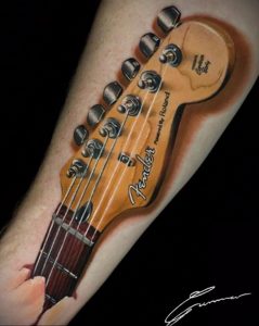 Реалистичная цветная татуировка гитарного грифа Fender на предплечье. Изображение выглядит как фотография.