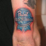 Фото минималистичной татуировки на руке с изображением логотипа Pabst Blue Ribbon. Простой и лаконичный рисунок татуировки в синих и красных тонах.