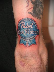 Фото минималистичной татуировки на руке с изображением логотипа Pabst Blue Ribbon. Простой и лаконичный рисунок татуировки в синих и красных тонах.
