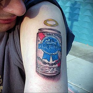 Фото татуировки на плече с изображением банки пива Pabst Blue Ribbon и золотого нимба над ней. Детализированный рисунок татуировки с юмористическим подтекстом.