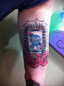 Фото татуировки на бедре с изображением банки пива Pabst Blue Ribbon, украшенной красными розами и лучами света. Детализированный рисунок татуировки в стиле олдскул.