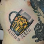 Фото тату на боку: кружка пива с пеной и надписью 'FLUCH & SEGEN'. Реалистичный рисунок татуировки.