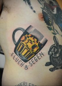 Фото тату на боку: кружка пива с пеной и надписью 'FLUCH & SEGEN'. Реалистичный рисунок татуировки.